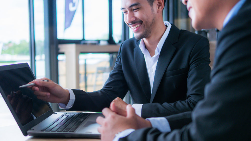 Photo of a man in a suit. He's smiling while pointing at a laptop screen while another man in a suit watches.