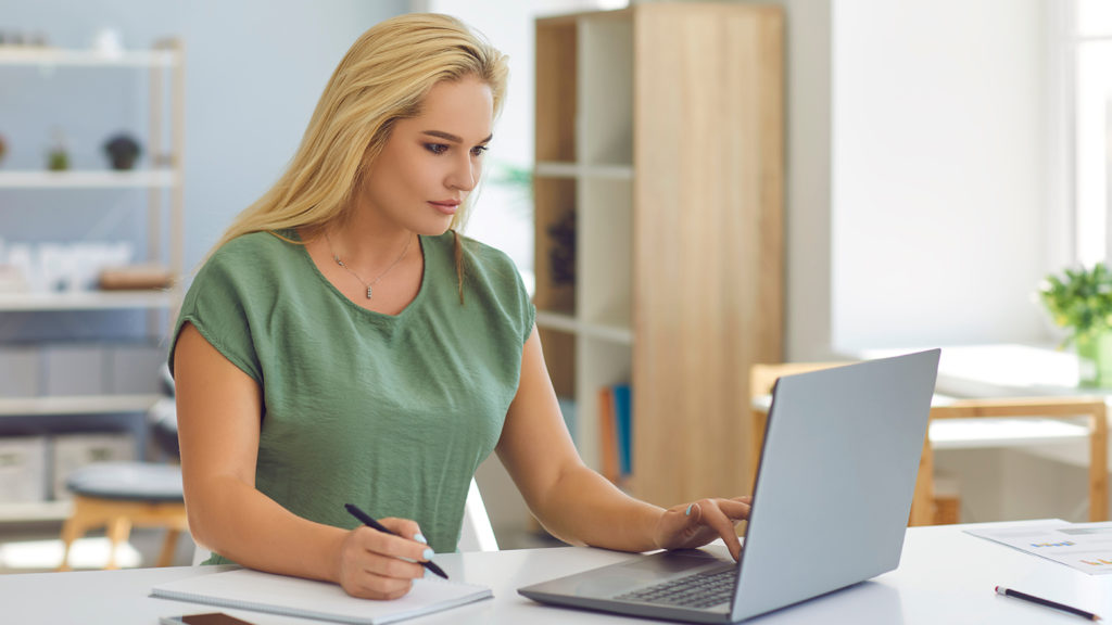 A woman sitting at a desk. She is using a laptop computer and writing on a notepad.